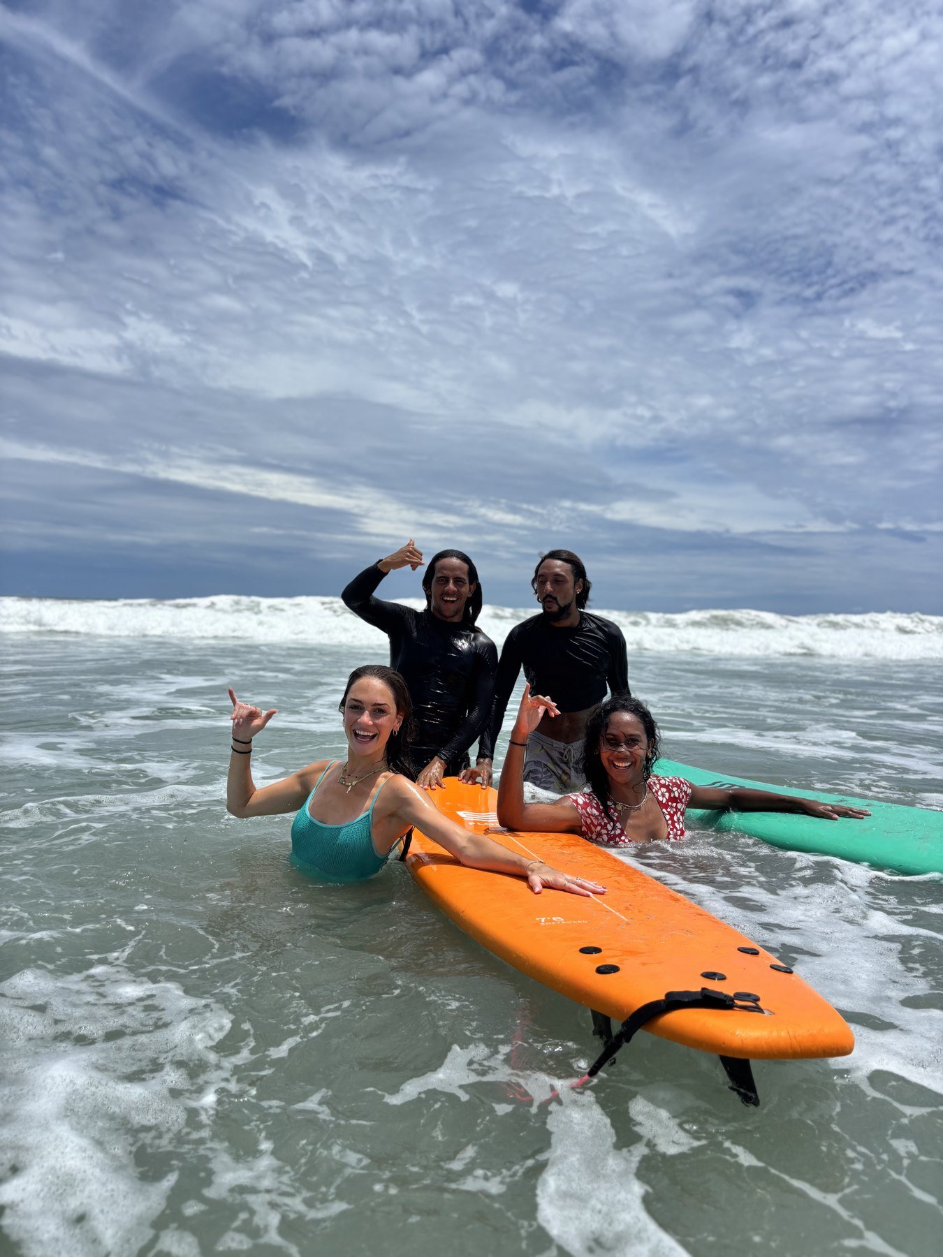 A surf lesson group throwing shakas around an orange soft board in the whitewater